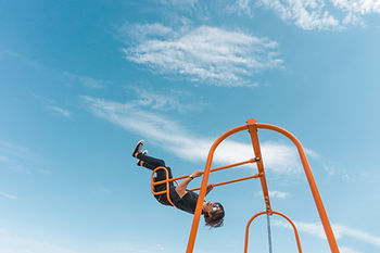 Person joyfully swinging upside down on orange playground bars under a bright blue sky with light clouds.