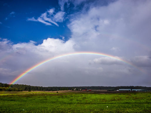 A vivid rainbow arches over a lush green field, set against a blue sky with white clouds. A red barn is visible in the distance.