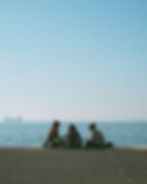 Three people sitting on a boardwalk by the sea, talking. Clear blue sky, calm ocean in background. Cargo ship in the distance. Connection.