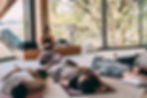 Meditation instructor holds a singing bowl while students lie on mats in a bright room with large windows and trees outside.