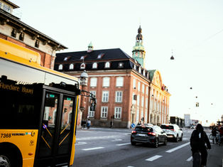 Yellow bus on city street with cyclists and cars. Historic brick building in the background. Traffic lights and evening light create a calm mood.