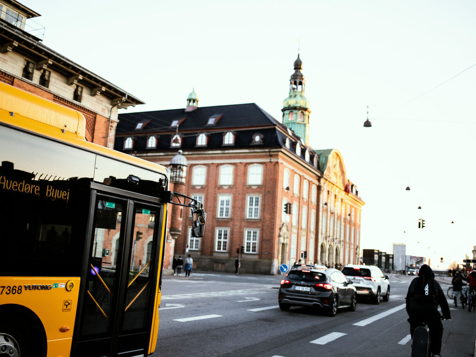 Yellow bus on city street with cyclists and cars. Historic brick building in the background. Traffic lights and evening light create a calm mood.