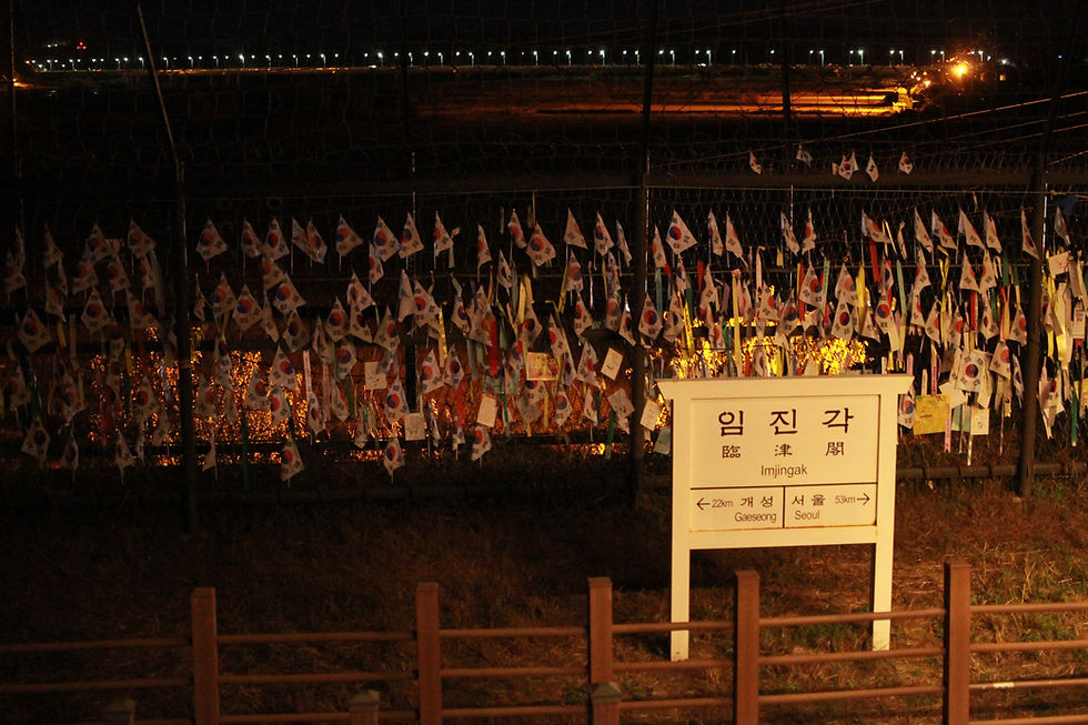 Korean flags hang on a fence under night lights at Imjingak. A sign displays distances to Gaeseong and Seoul, set against a dark sky.