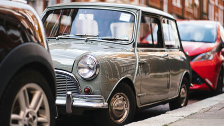 Voiture Mini vintage grise garée dans une rue pavée, avec une voiture rouge floue derrière, ambiance urbaine calme.