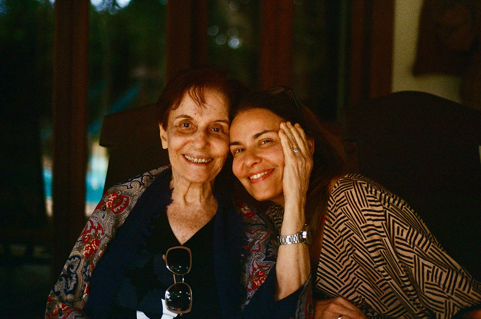Two smiling women sit close together indoors, one resting her hand on the other's face, in patterned shawls.