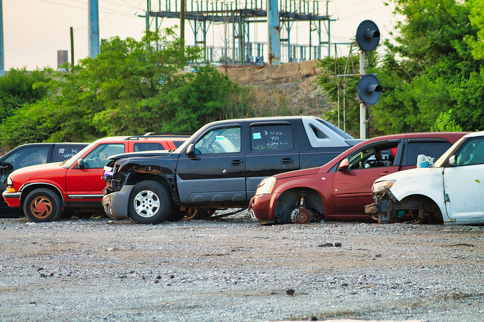 Damaged cars parked on gravel, missing wheels, surrounded by greenery and industrial structures. Notes on windows, late afternoon light.