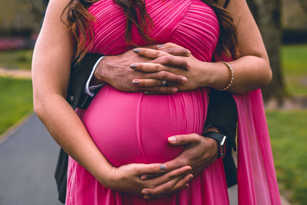 Expecting mother wearing a pink dress, with her partner holding her belly from behind, wearing a black suit. Captured by a maternity photographer in New York.