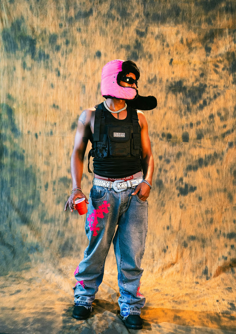 Person wearing a pink helmet, black vest, and jeans with pink designs, holding a red cup. Text "CALVIN KLEIN" visible. Earthy backdrop.