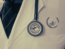 Close-up of a doctor's white coat with a Littmann stethoscope on the chest pocket. A striped tie is visible. Emblem and buttons add detail.