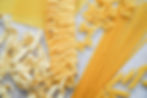 Assorted uncooked pasta varieties including spaghetti, fettuccine, farfalle, and penne are displayed on a textured grey background.