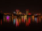 Stadtpanorama bei Nacht mit beleuchteten Hochhäusern und bunten Lichtern, die sich im ruhigen Wasser spiegeln; stimmungsvolle Atmosphäre.