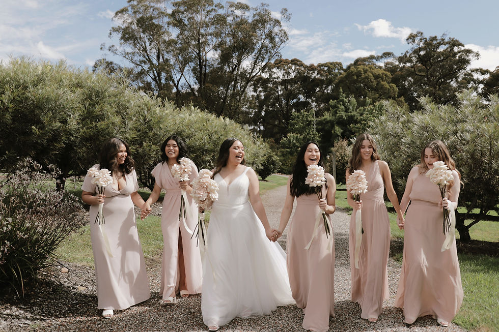 Bride in white and five bridesmaids in pink holding flowers walk joyfully through a garden path, surrounded by greenery and trees.