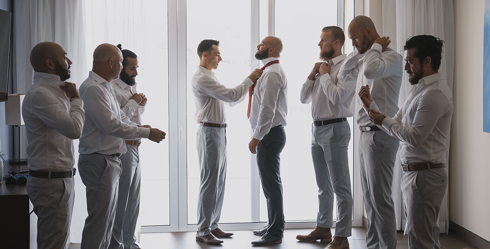 Groomsman fixing the tie of the groom standing by the window while the other six groomsmen watch while fixing their own attire.