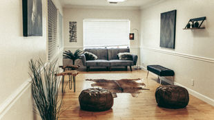 Cozy living room with a gray sofa, fur pillows, cowhide rug, and poufs on a wooden floor. Art decorates walls; a lamp softly glows.