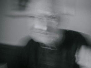 Blurred image of an elderly person in grayscale, sitting indoors. The motion creates a ghostly effect, with soft lighting and indistinct background.