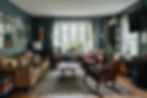 Cozy living room with leather couch, armchairs, plants by window, patterned rug, and wall art. A "TATTOO" sign hangs on a dark teal wall.