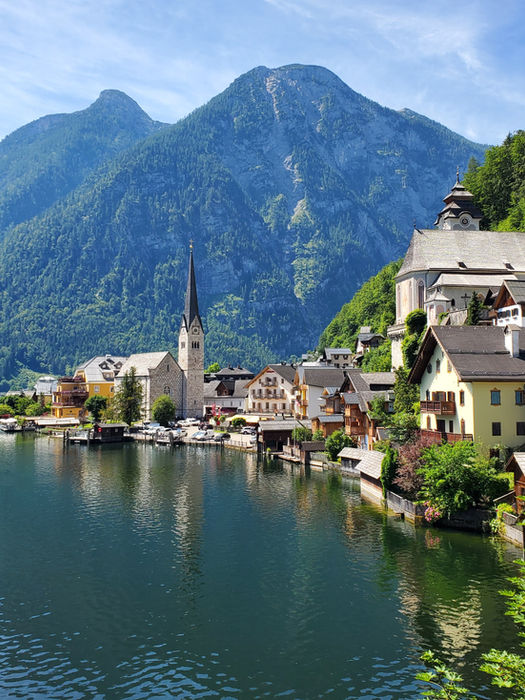 A picturesque view of Hallstatt, Austria, with traditional alpine houses nestled between a serene lake and majestic, tree-covered mountains under an overcast sky. A prominent church steeple rises in the center of the village.