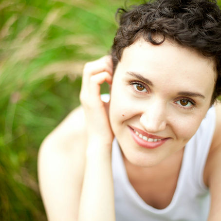 Smiling individual wearing a white top sits in green grass, looking up with a joyful expression. Bright, natural setting.