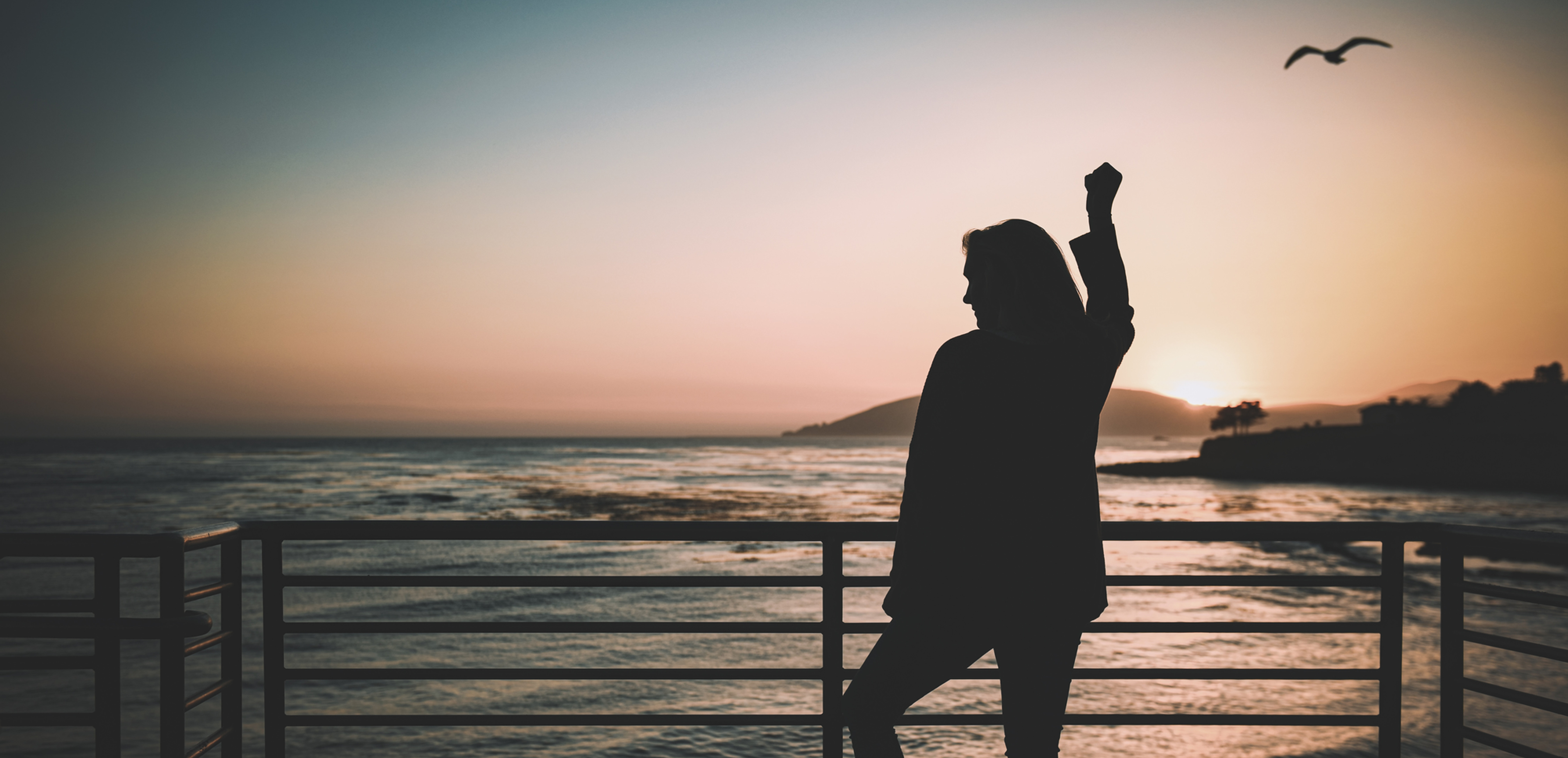Silhouette of a woman raising one arm on a seaside railing at sunset, with a bird overhead and calm ocean beyond.