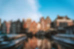 Canal with docked boats reflecting colorful, historic buildings in a sunlit, vibrant setting. Dutch text visible on a building facade.