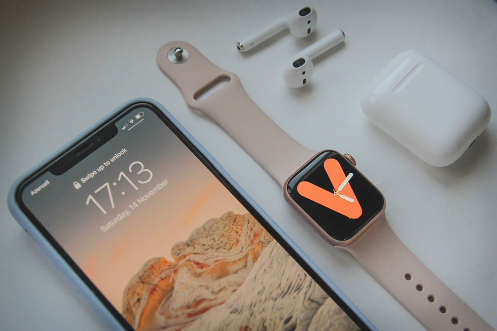 A smart phone, digital watch, wireless headphones, and a headphones case laying side by side on a table