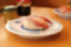 Two pieces of sushi on a white plate with "Kura" text. Wasabi jar and soy sauce in the background. Warm, inviting restaurant setting.