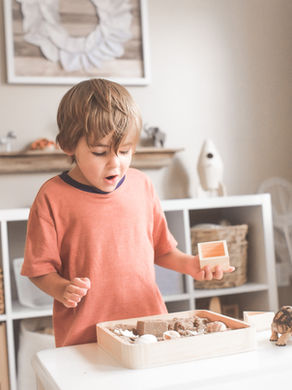 Child in orange shirt and surprised expression, playing with a wooden tray of sand and shells on a table. Home setting with shelves behind.