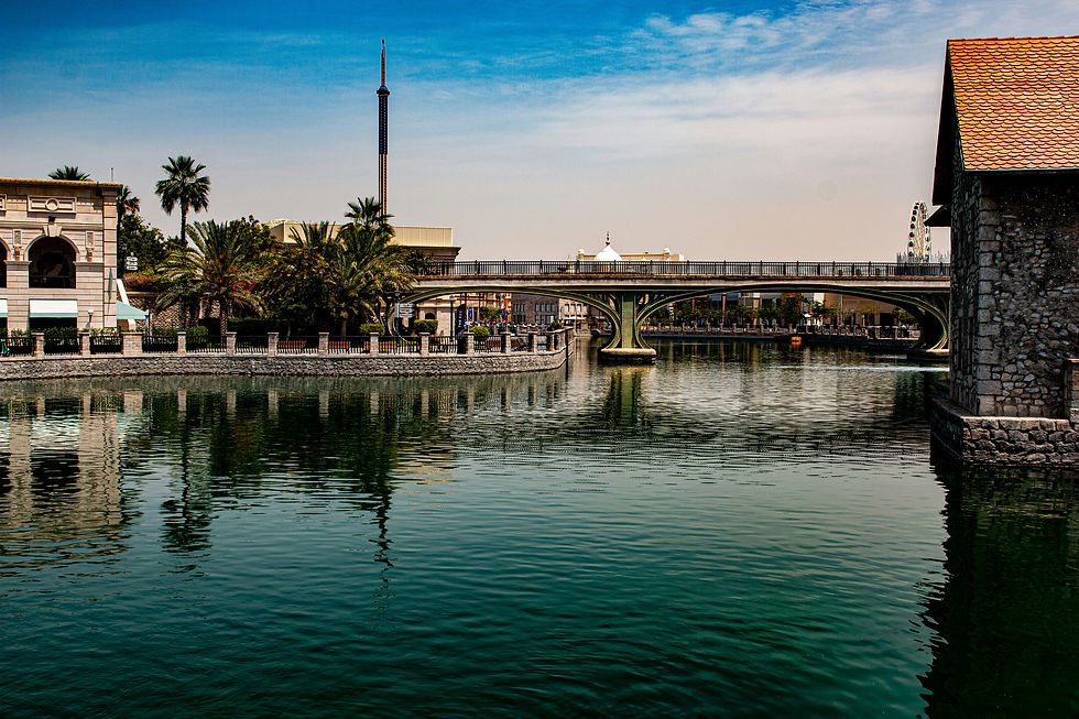 Calm river scene with a stone bridge, palm trees, and buildings, under a clear blue sky. Reflections ripple on the water's surface.