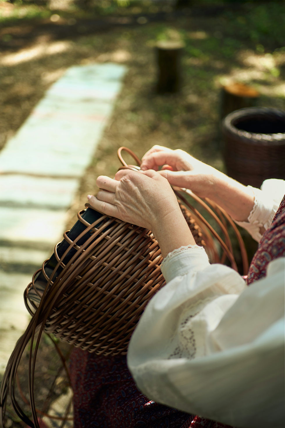 Hands weaving a wicker basket outdoors in warm sunlight, with a blurred garden path and wooden stumps in the background.