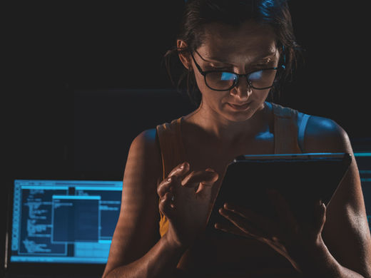 Woman with glasses taps a tablet in a dark room, lit by blue code-filled computer screens, focused and intent.