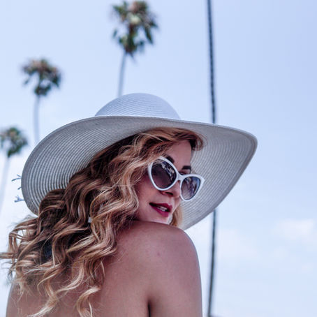 Woman with wavy hair wears a wide-brimmed hat and sunglasses, with palm trees in the background against a clear blue sky.