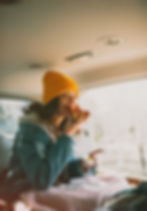 A woman in a yellow beanie enjoys pizza inside a car. She wears a denim jacket, smiling with sunlight streaming through the window.