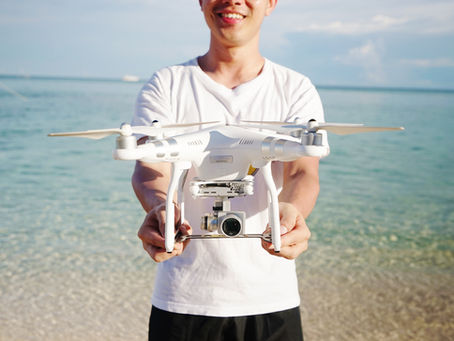 Smiling man holding white drone at beach. Clear blue water and sky in background, wearing a white shirt and black shorts.