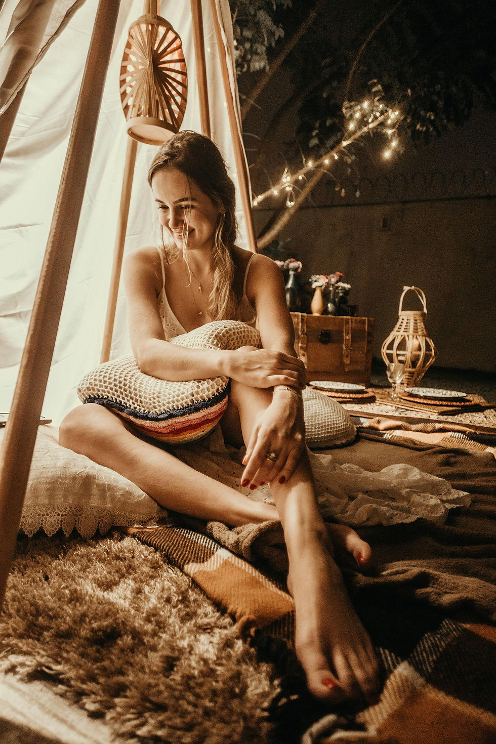 Smiling woman sits cross-legged in a cozy boho tent, holding a striped pillow amid warm string lights and woven decor.