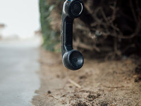 Hanging black telephone receiver over sandy ground and blurred background of shrubs, creating a mysterious and abandoned mood.