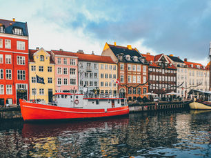 A scenic view of Nyhavn in Copenhagen, Denmark. A large red and white boat is moored in the canal, with a row of iconic, colorful 17th and 18th-century townhouses lining the waterfront under a cloudy sky.