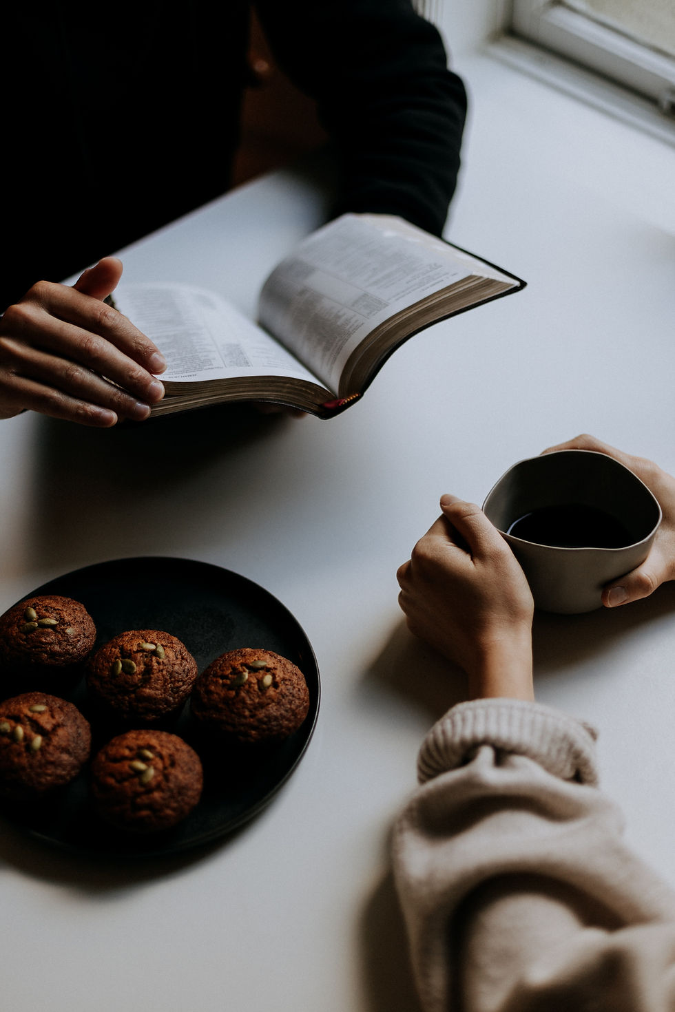 Two pairs of hands are seen resting at a table as two people sit across from each other with a dark plate of muffins next to them. One is flipping through a Bible in their hands white thebother has a cup with a dark beverage.