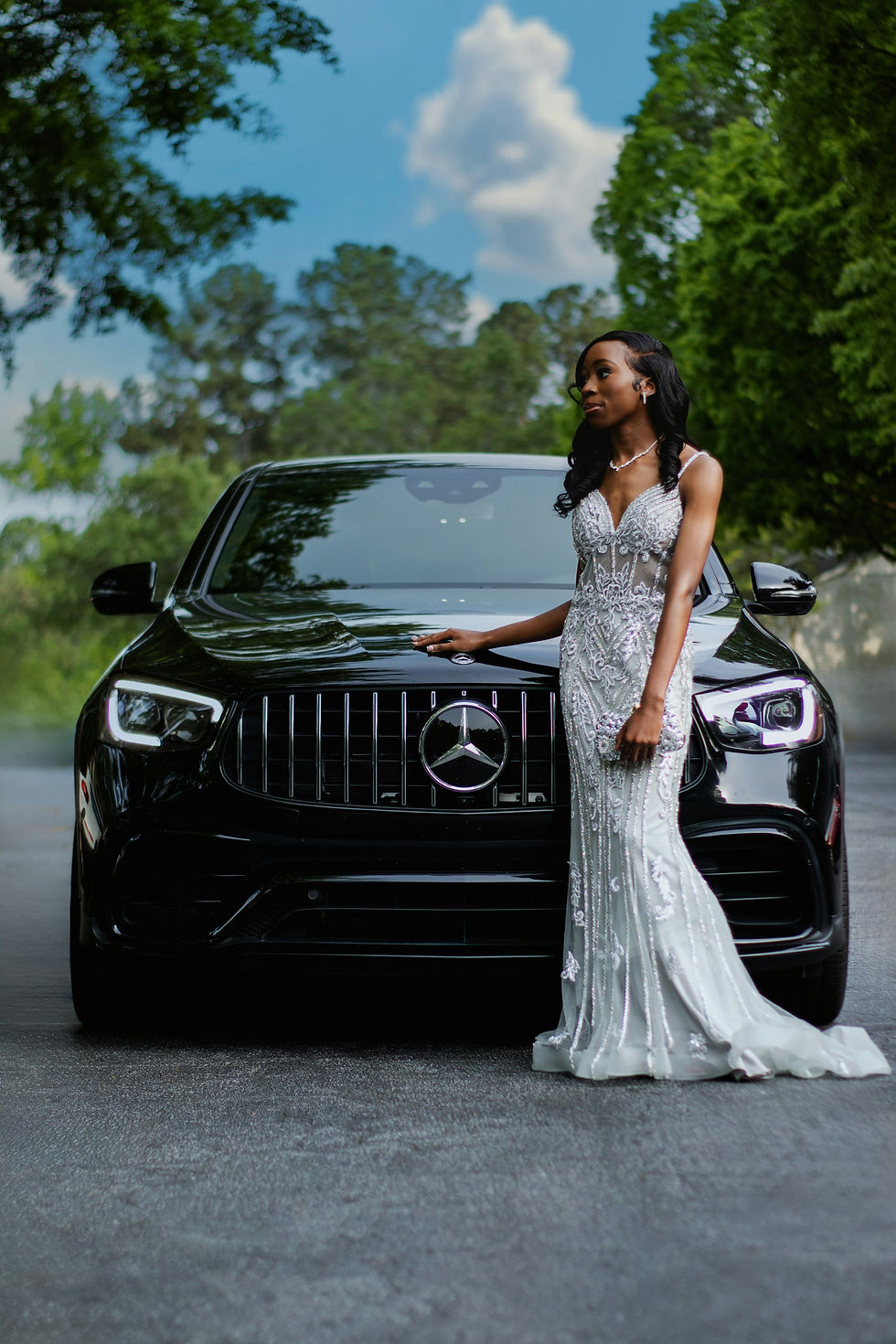 A woman in a shimmering evening gown stands confidently beside a sleek Mercedes Benz, exuding style and sophistication.