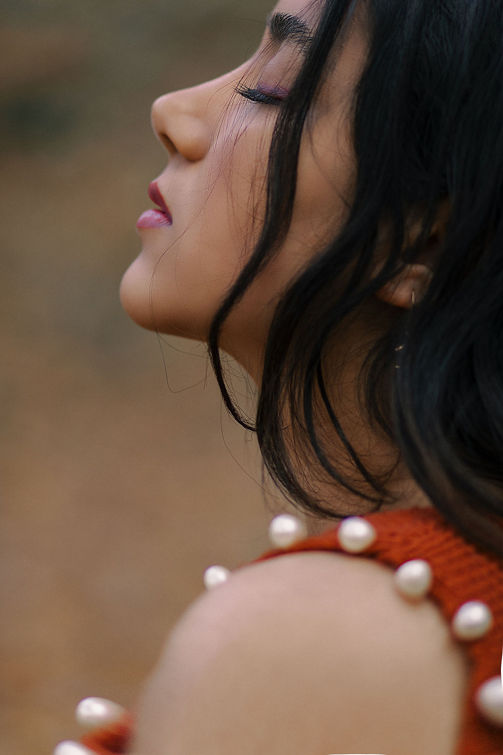 Profile of a woman with closed eyes, long dark hair, wearing a pearl-studded orange top. Background is blurred, creating a serene mood.