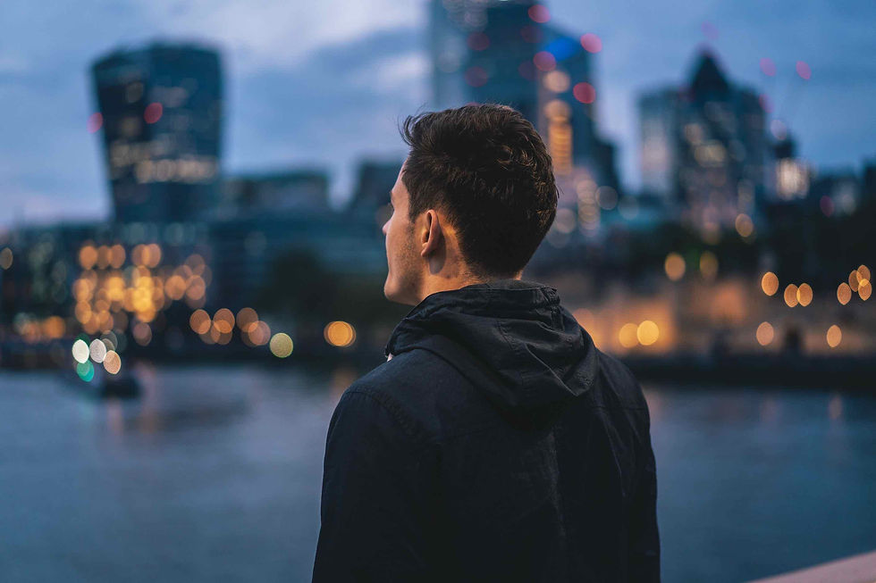 Man in black jacket gazes at city skyline at dusk, blurred lights reflecting in water, creating a serene, contemplative mood.