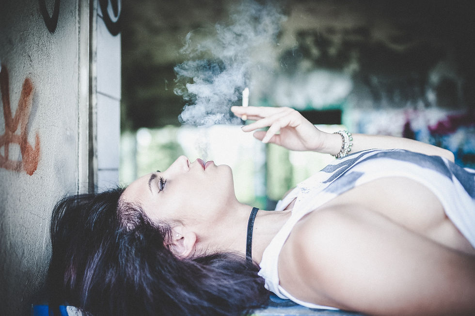 Attractive Caucasian young woman with black hair in profile, lying down next to wall smoking a cigarette.