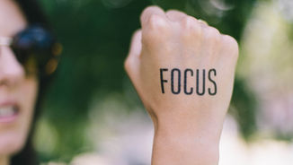 Close-up of a fist with "FOCUS" written on it, blurred person in sunglasses in the background, outdoors with greenery.