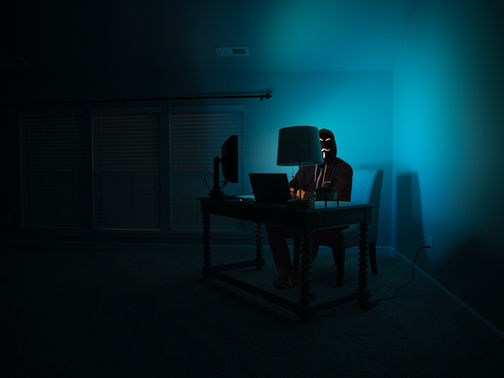 Hooded person wearing a Guy Fawkes mask works at a desk with laptop and monitor in a dark blue-lit room.