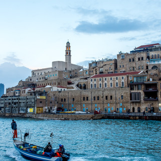 A traditional blue wooden fishing boat in the calm waters of the ancient Jaffa Port, with the Old City walls rising in the background under a soft blue twilight sky.