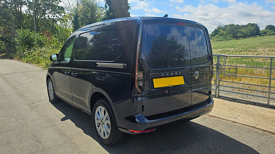 Rear three-quarter view of a dark blue or black commercial van parked on an outdoor driveway near country fencing and greenery