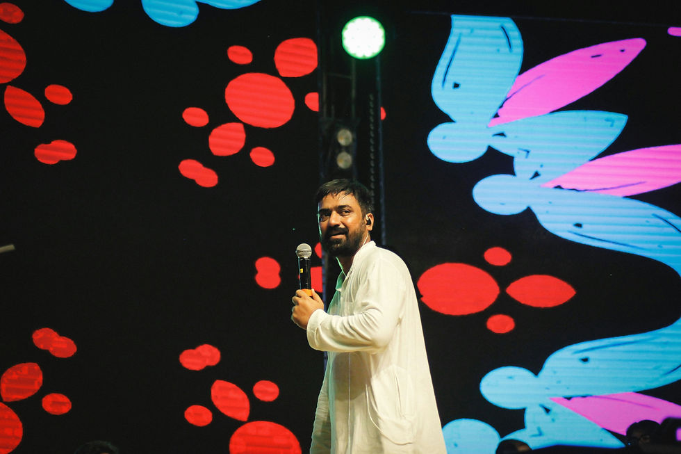 Man in white holding a mic on stage, vibrant LED backdrop with red and blue abstract patterns, bright and lively atmosphere.