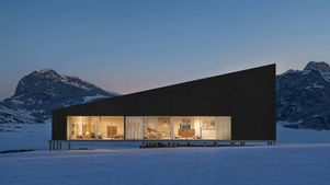 Modern black cabin glowing warmly in snowy mountains at dusk, with a clear blue sky and no people visible.