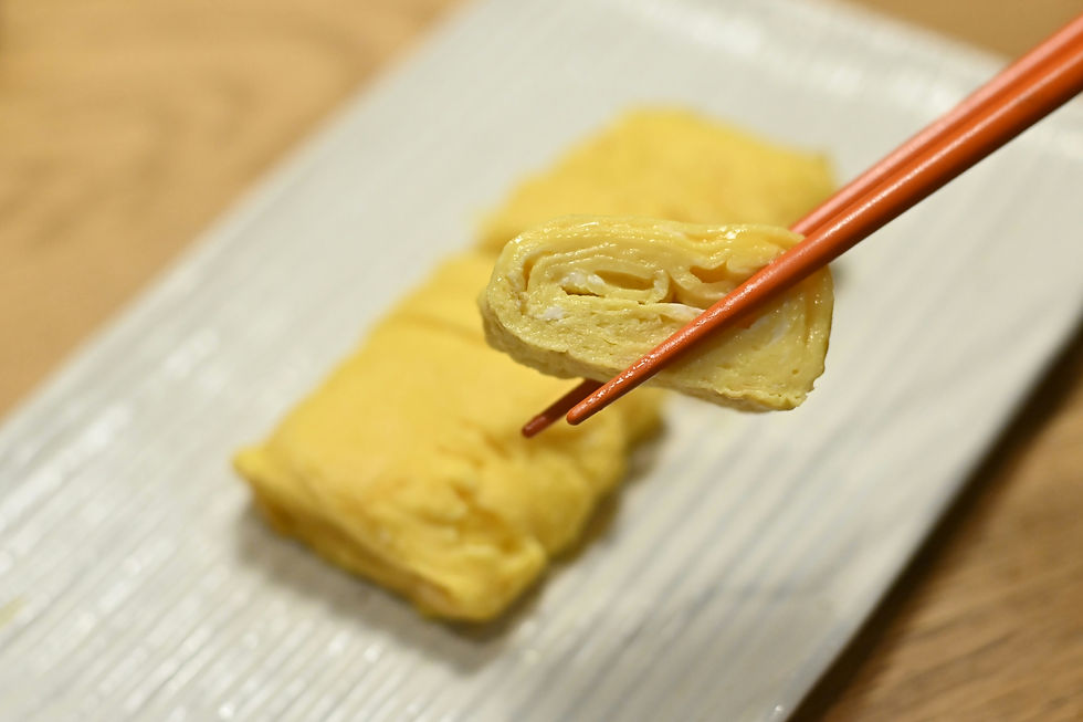 Chopsticks hold a piece of tamagoyaki above a white plate with more slices. The scene has a warm, inviting atmosphere with neutral tones.