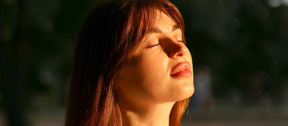 A woman taking a deep breath with eyes closed, looking up at sunset.