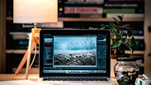 Laptop on a cozy desk shows Adobe Lightroom editing a frosty landscape; lamp, plant, books, and warm light frame the scene.